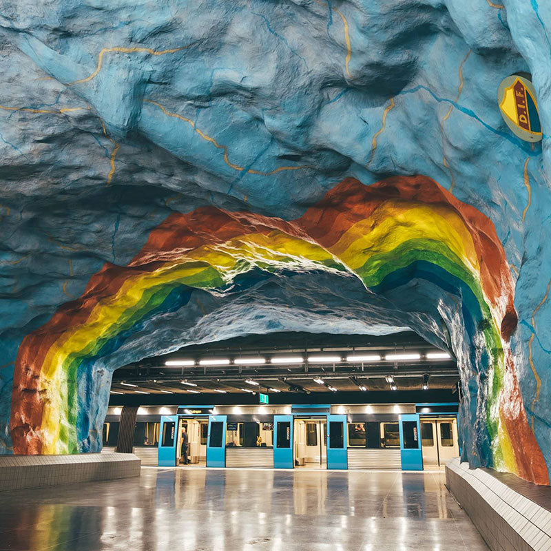 A subway platform seen through a rough blue cave-like arch painted with a rainbow, with a train waiting