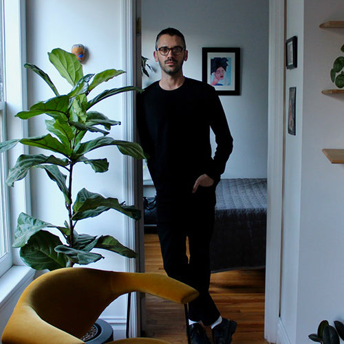 A mustard yellow curved armchair beside a fiddle leaf fig, with Conor Ford standing in a doorway to a bedroom