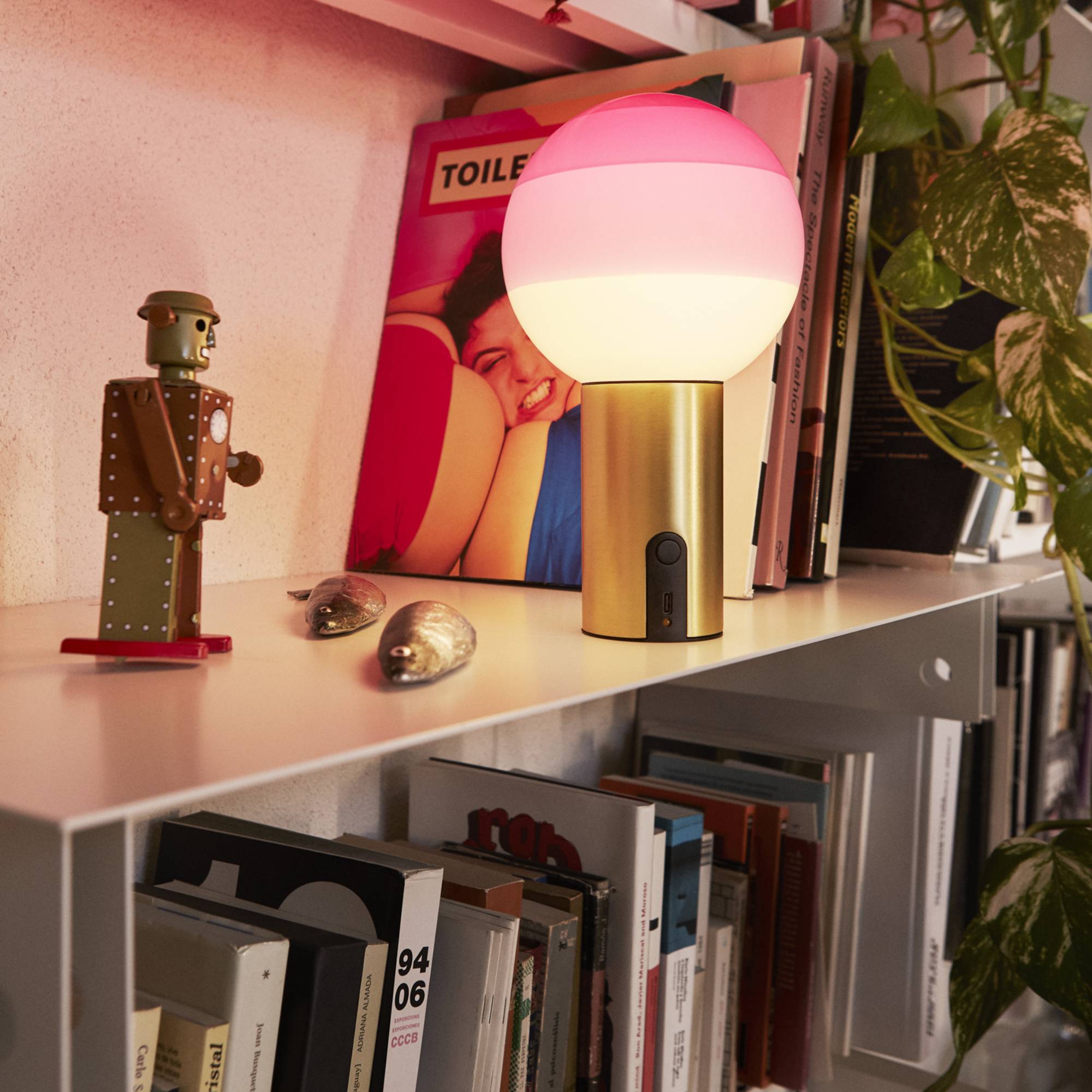 A cordless table lamp with a brass base and a pink-to-white glass globe on a white shelf by books