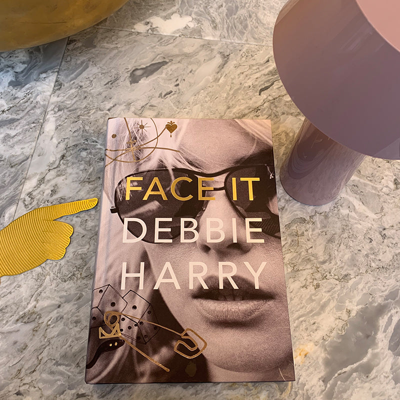 A hardback book with a person's portrait on its cover, lying on grey marble beside a lilac table lamp