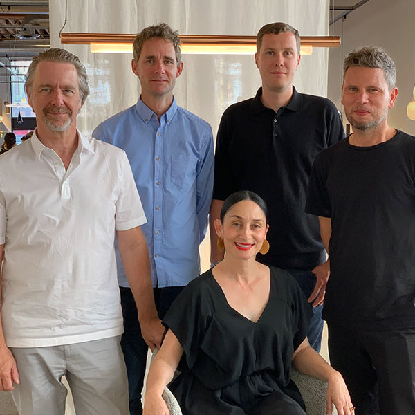 Five people posed together indoors, four standing and one seated, under a long glowing copper light