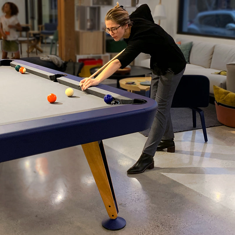 A pool table with a navy rail and angled wooden legs as a person leans over to take a shot in a lounge