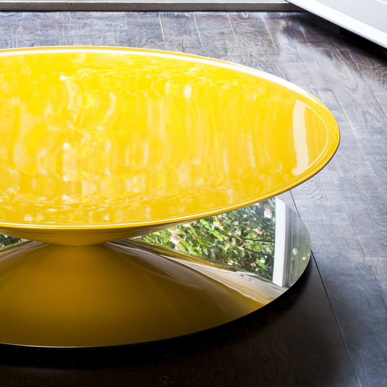 A glossy yellow oval coffee table on a mirrored conical base, on a dark wood floor