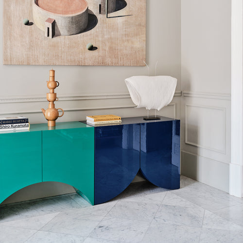 A glossy sideboard with teal and navy lacquered arched panels on a marble floor, below an abstract painting