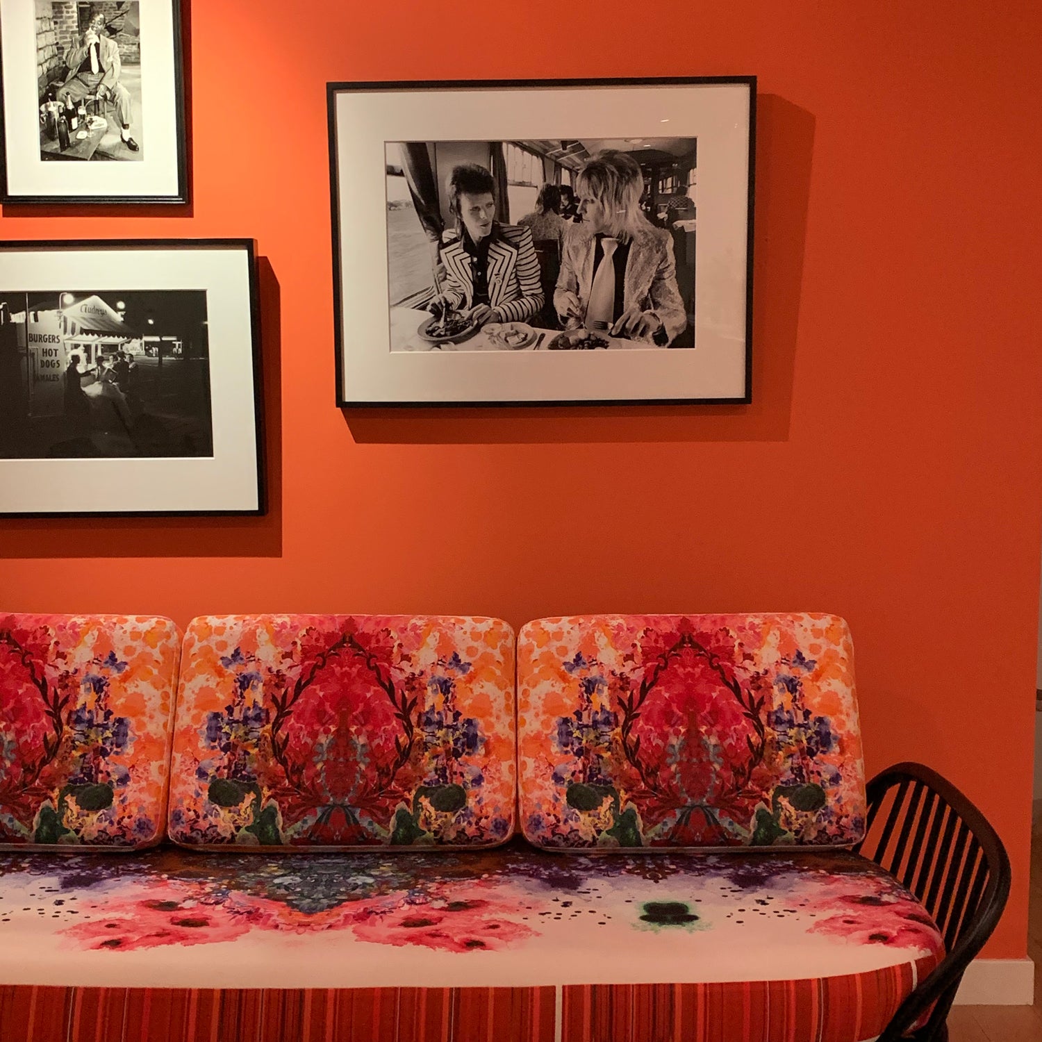 A sofa with black spindle arm and cushions in a bright floral pattern, below framed black-and-white photos on an orange wall