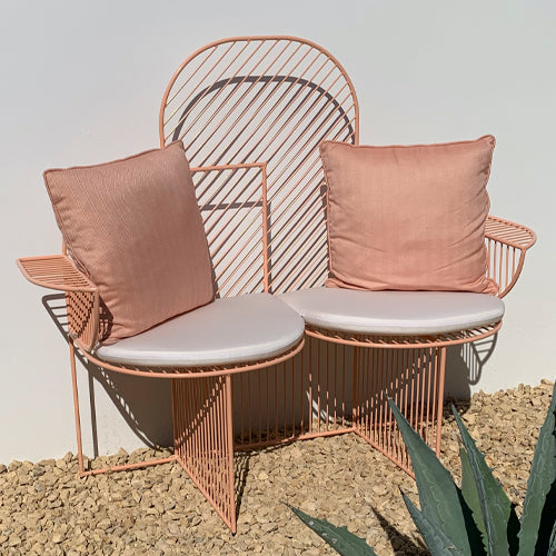A pink metal outdoor bench with two seats, arched slatted back and blush cushions, on gravel by a white wall