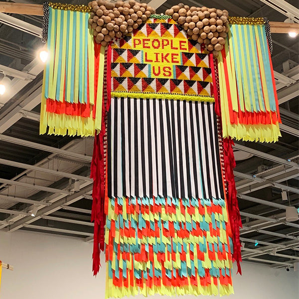 A large hanging garment of colourful ribbon fringe and wooden beads with beaded text reading People Like Us