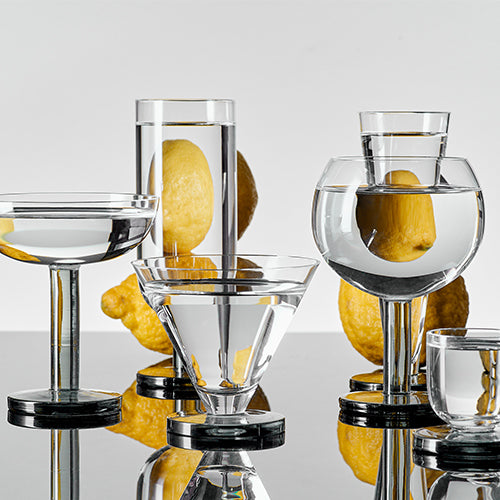A set of clear stemmed glasses with dark bases filled with water, magnifying whole lemons behind them