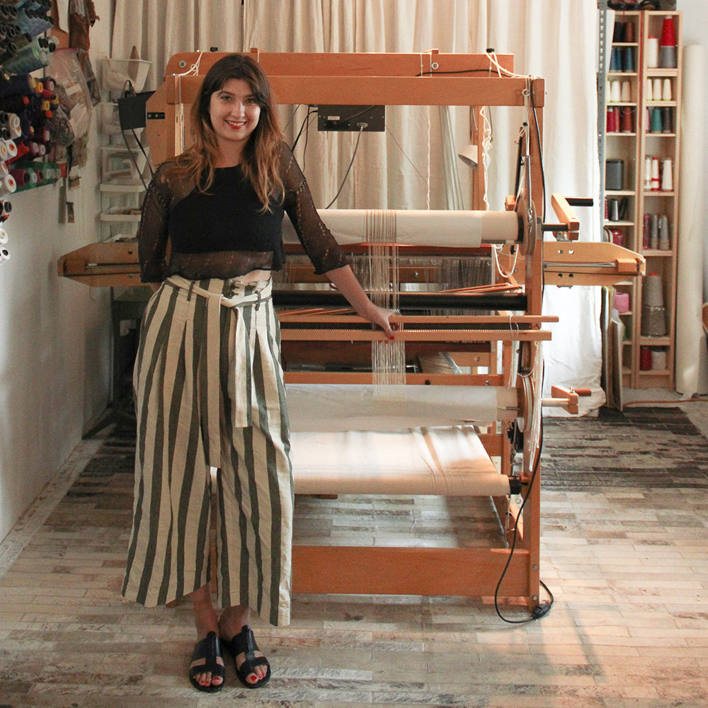 Iris Murphy standing beside a large wooden floor loom in a studio, with spools of thread on shelves behind