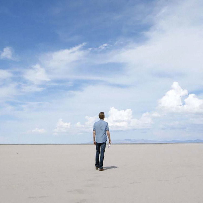 A person seen from behind walks across a vast pale salt flat under a wide blue sky with scattered clouds