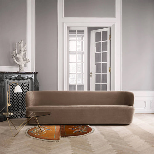 A long curved-back sofa in taupe velvet in a pale room with herringbone floors and a glass side table