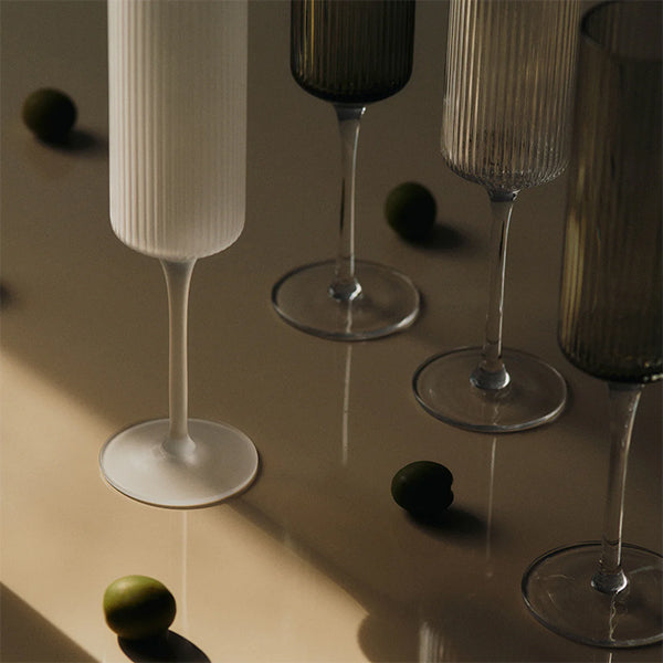 Four ribbed champagne flutes in frosted white, smoky grey and clear glass on a tan surface