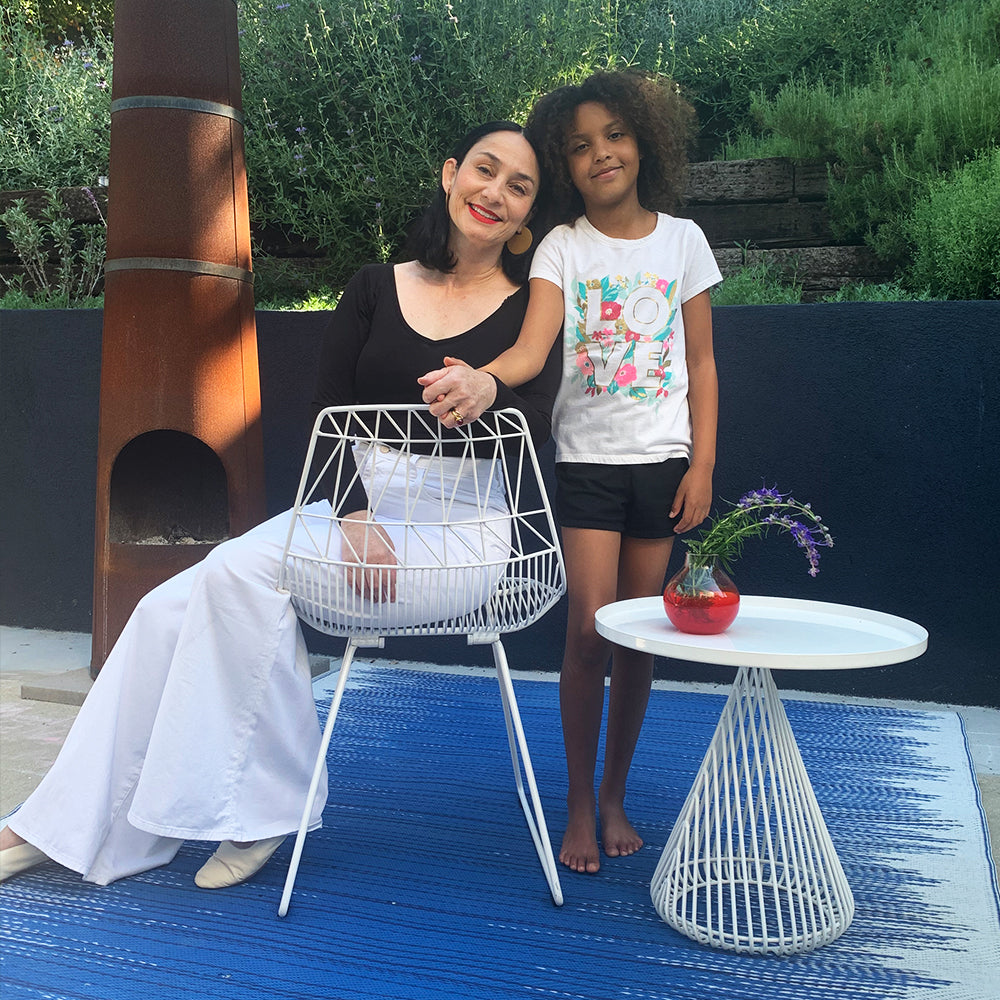 A white wire chair and round side table on a blue rug, with a seated person and a child standing beside them