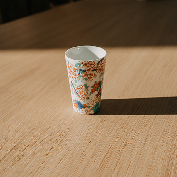 A white porcelain tumbler painted with orange blossoms and blue-green leaves on a light oak table in sunlight