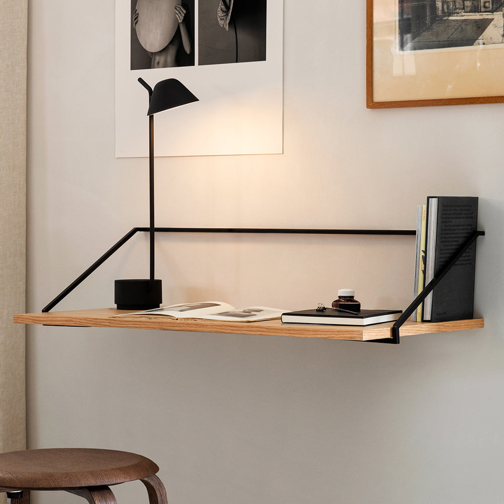 A wall-mounted light oak desk with thin black steel supports, a black desk lamp and an open book