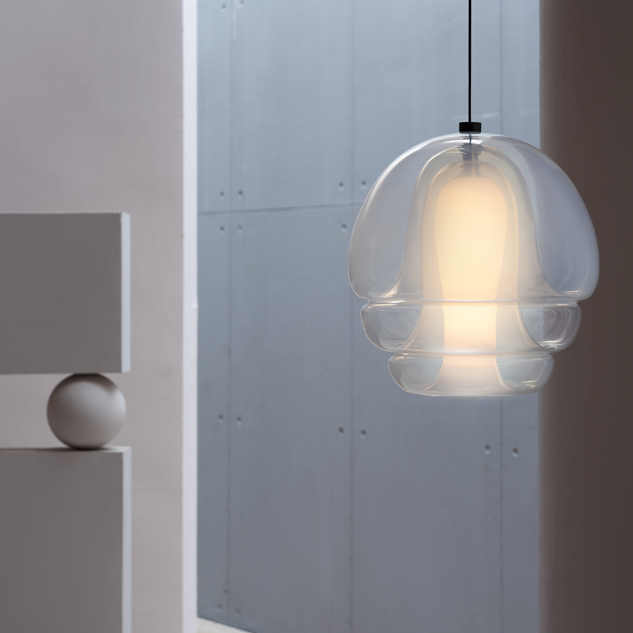 A glowing glass pendant with clear layered tiers around a frosted core, hanging against a grey concrete wall