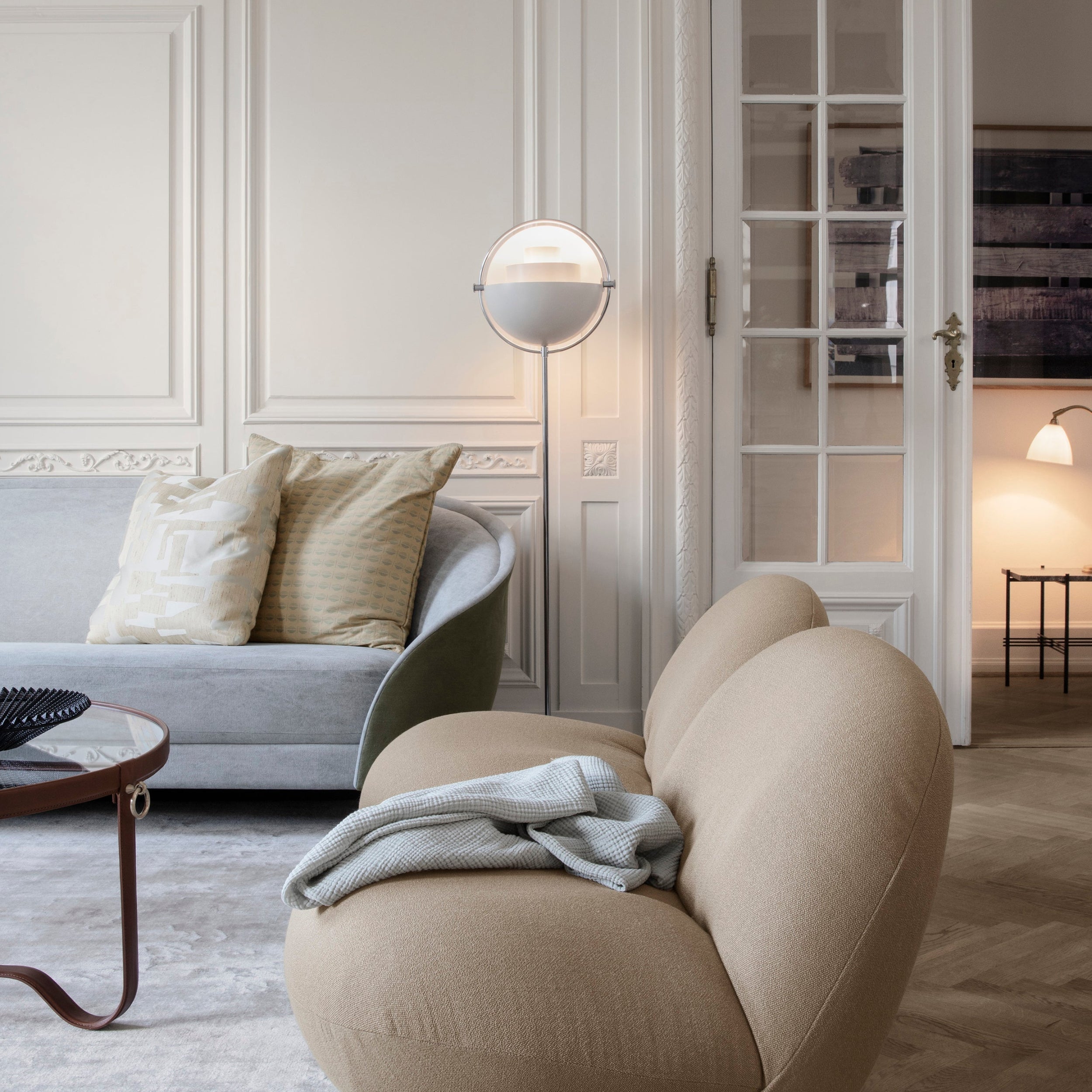 A round white floor lamp with a half-dome shade glows beside a pale grey sofa and a rounded beige armchair