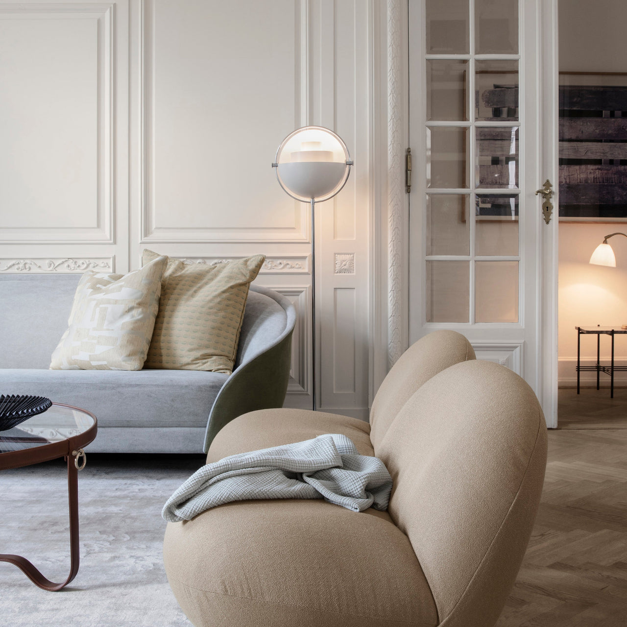 A round white floor lamp with a half-dome shade glows beside a pale grey sofa and a rounded beige armchair