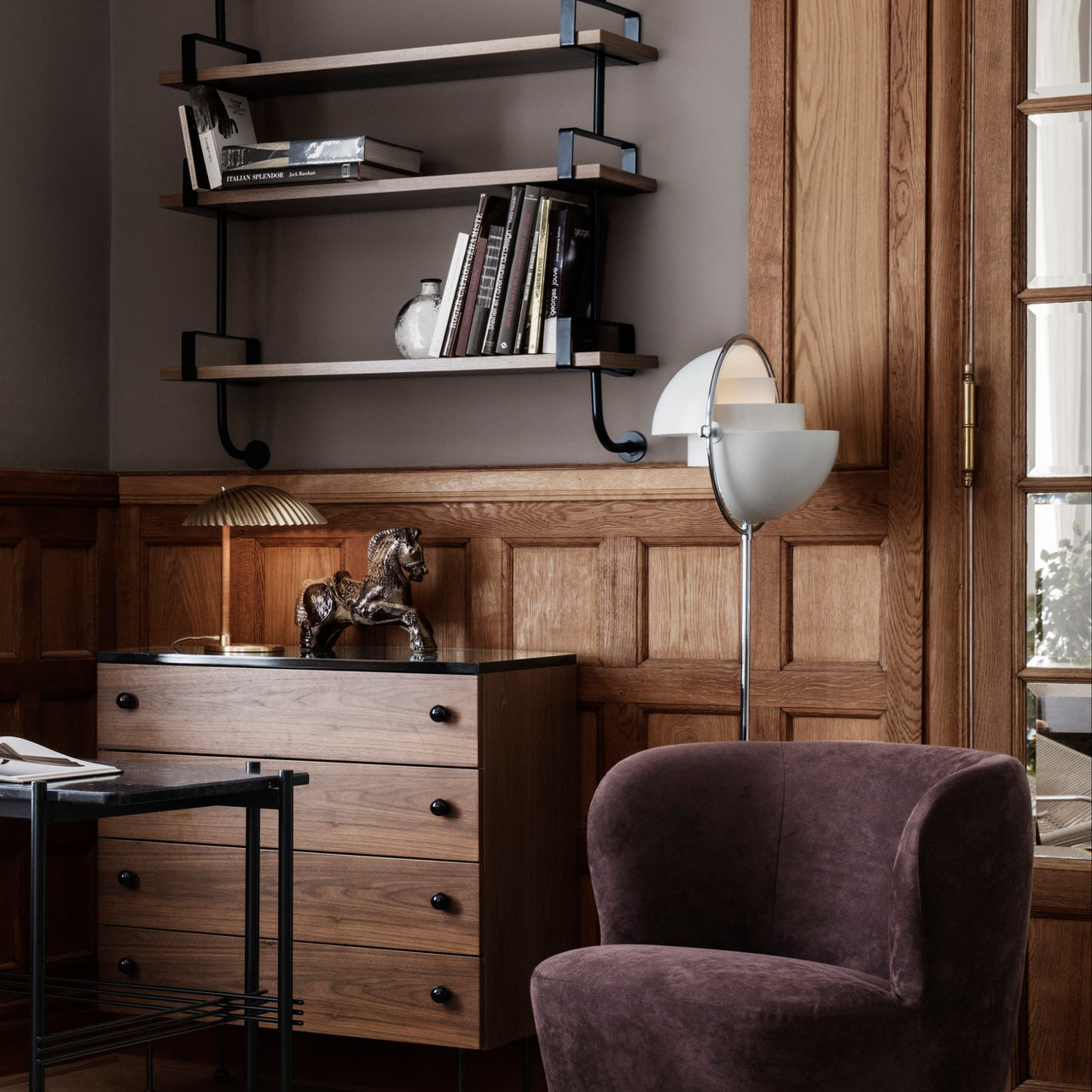 A white and brass floor lamp with a tilting half-dome shade beside a plum armchair and wooden dresser