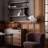 A white and brass floor lamp with a tilting half-dome shade beside a plum armchair and wooden dresser