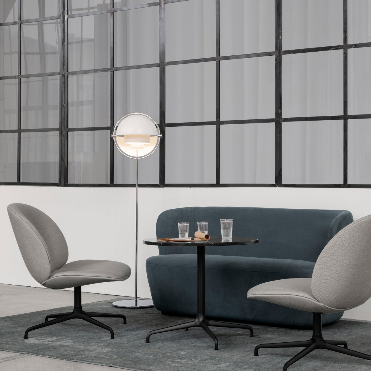 A floor lamp with a white round shade beside a blue-grey sofa, small round table and two grey swivel chairs