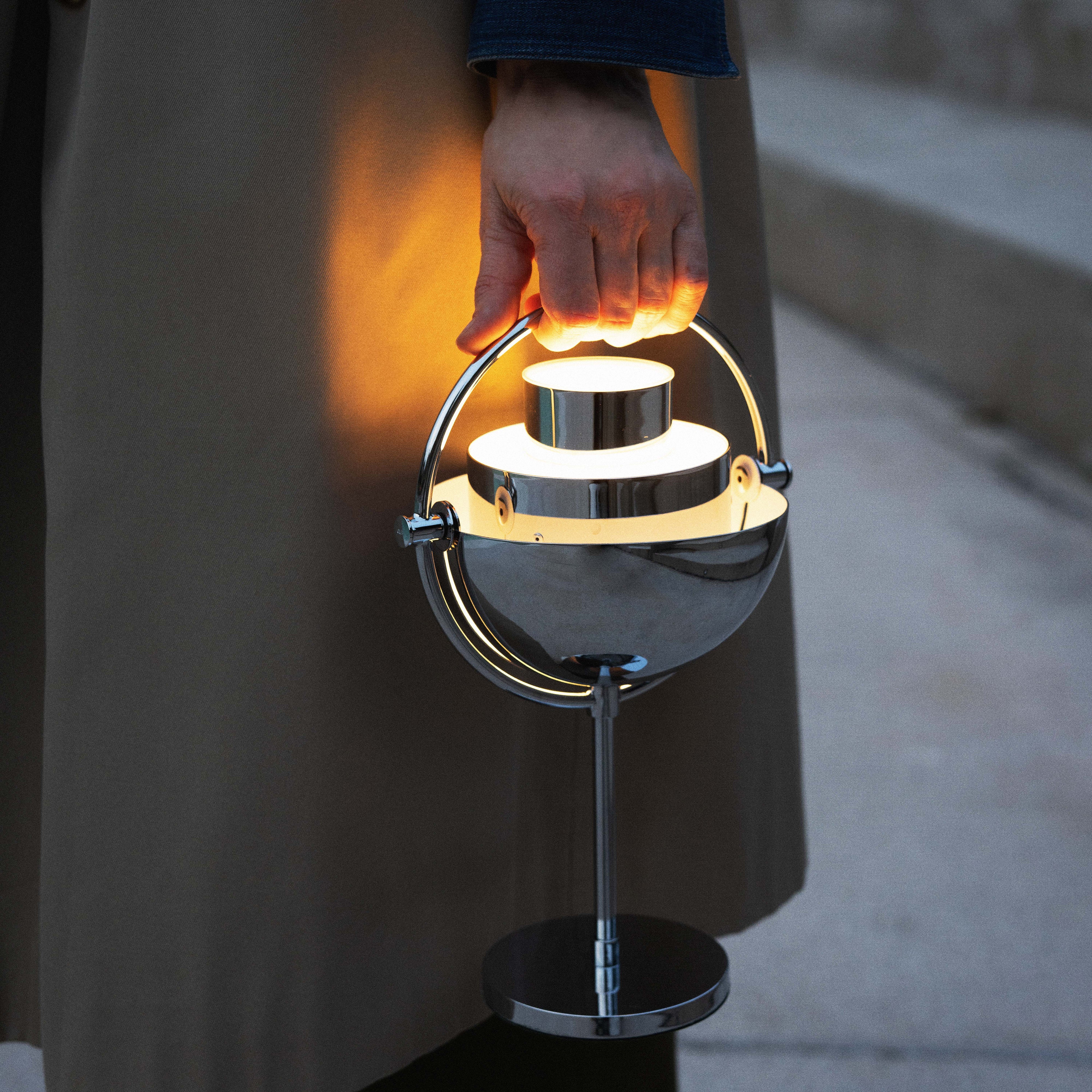 A glowing chrome portable lamp with a half-dome shade and round base, carried by its curved handle outdoors