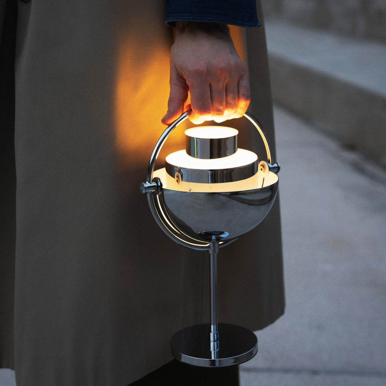 A glowing chrome portable lamp with a half-dome shade and round base, carried by its curved handle outdoors