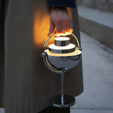 A glowing chrome portable lamp with a half-dome shade and round base, carried by its curved handle outdoors