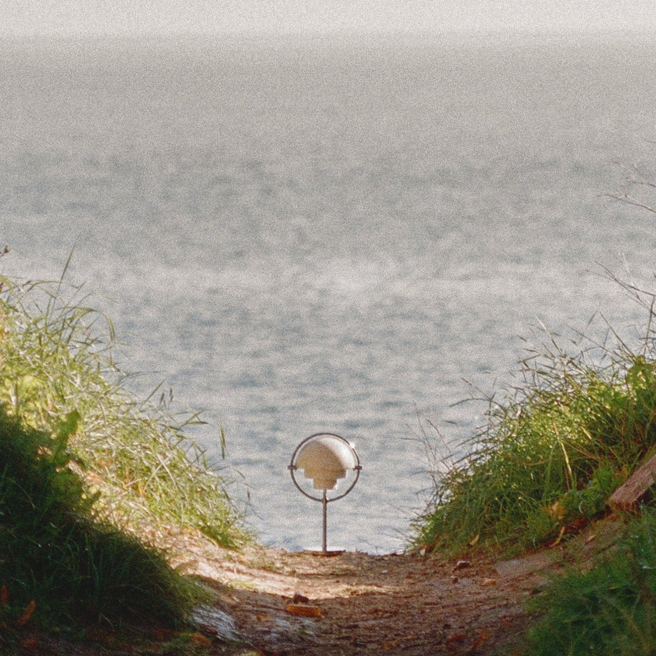A small portable lamp with a cream stepped shade in a thin ring stand, on a path overlooking the sea