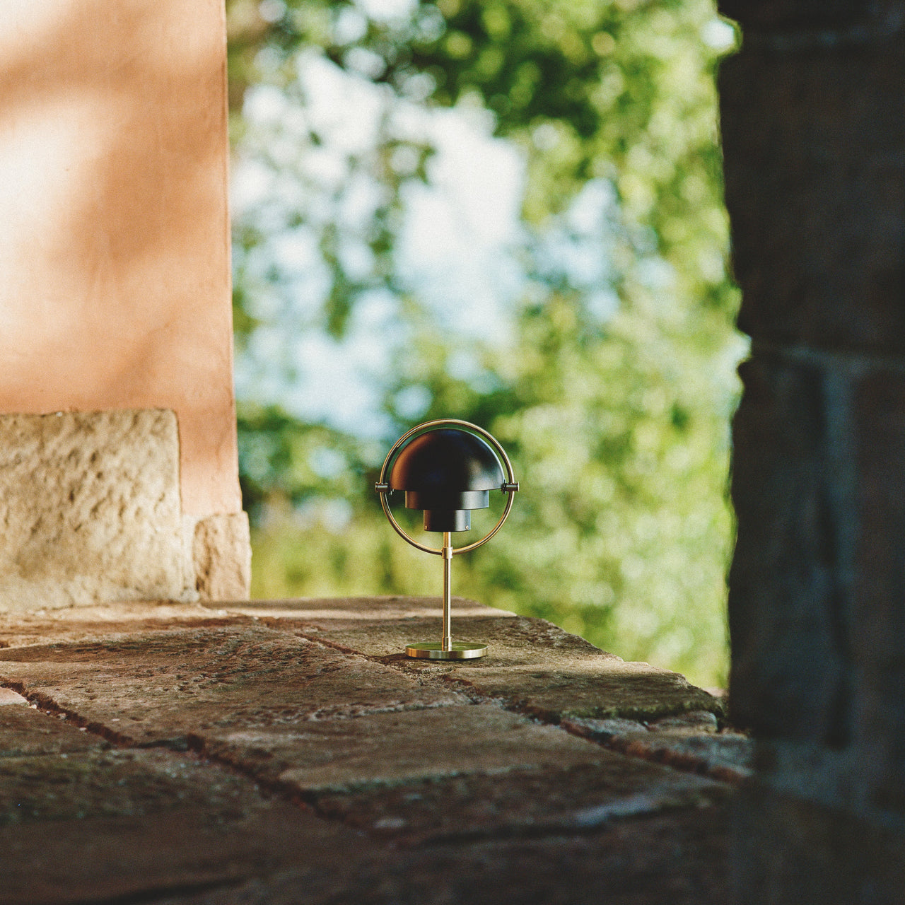 A small portable lamp with a black domed shade and brass stand on a stone ledge outdoors