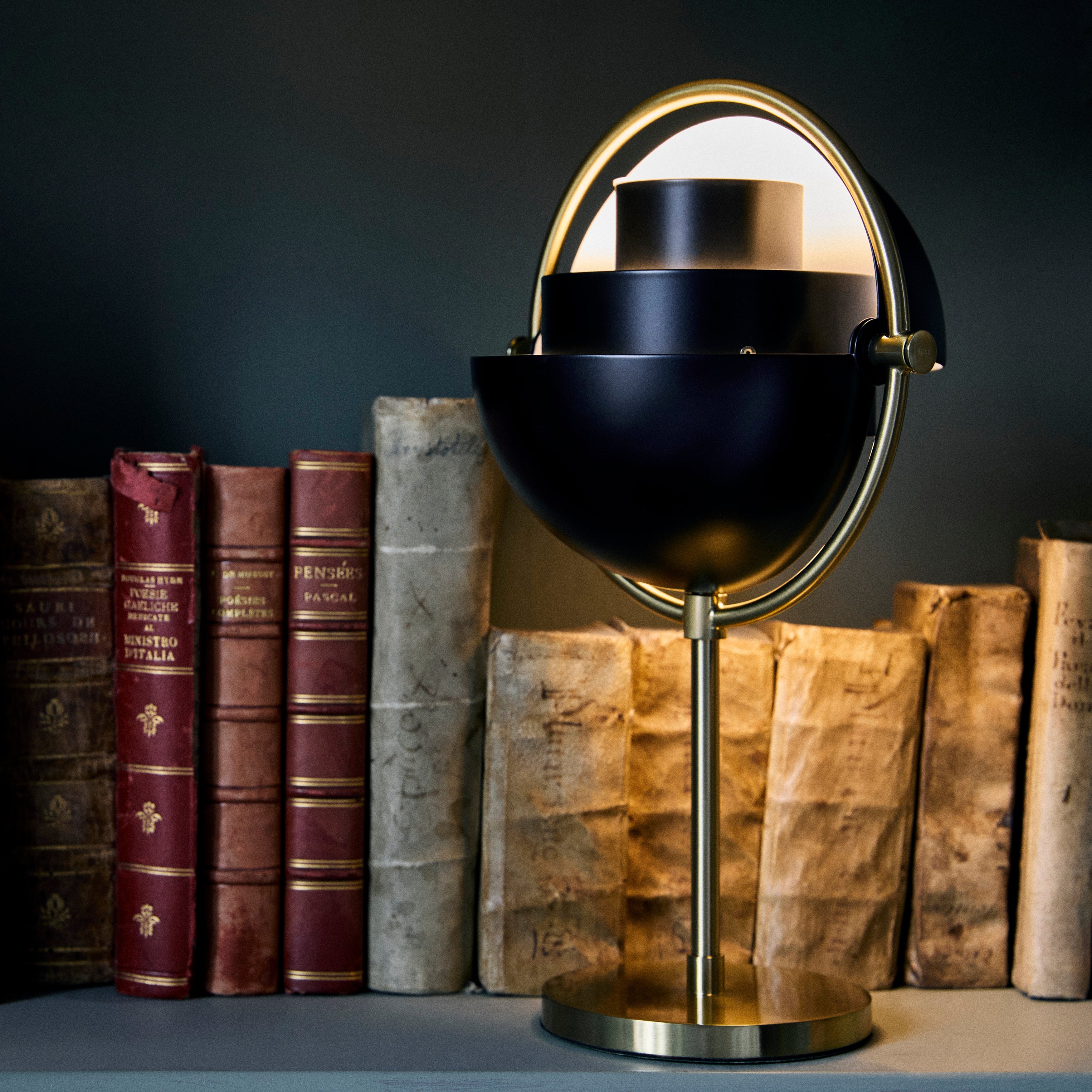 A black half-dome table lamp on a brass stem and round base, glowing in front of old books