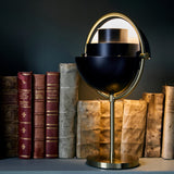 A black half-dome table lamp on a brass stem and round base, glowing in front of old books