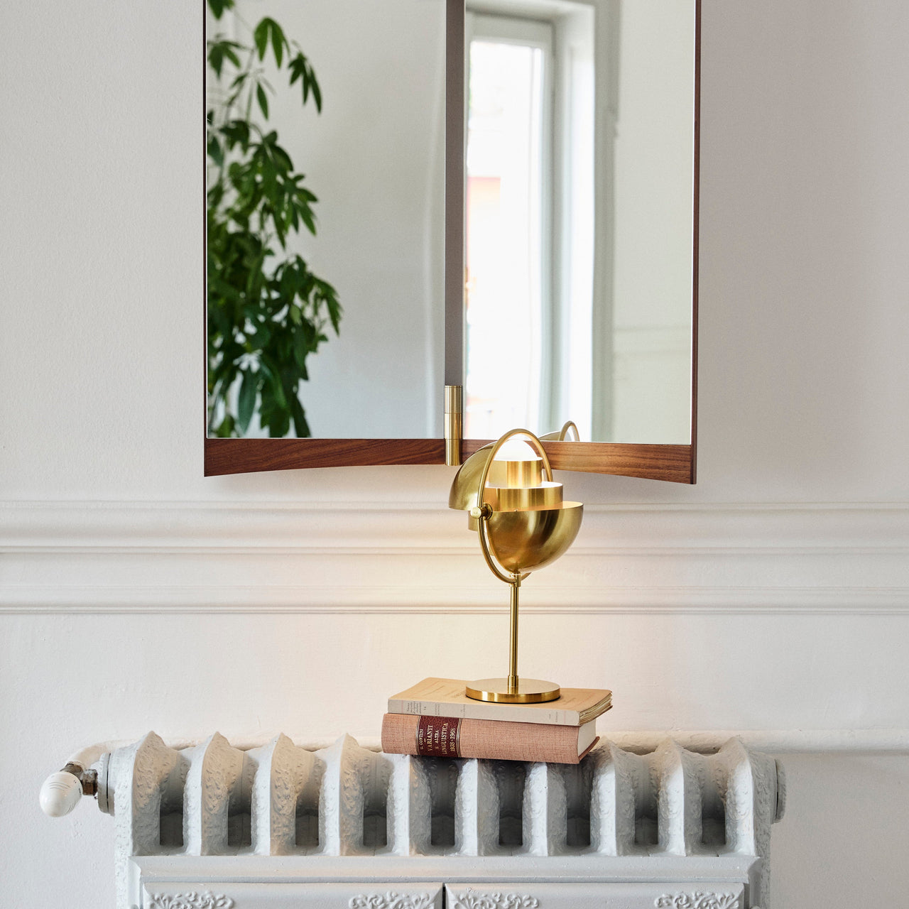 A polished brass table lamp with a round pivoting shade sits on two books beneath a wall mirror