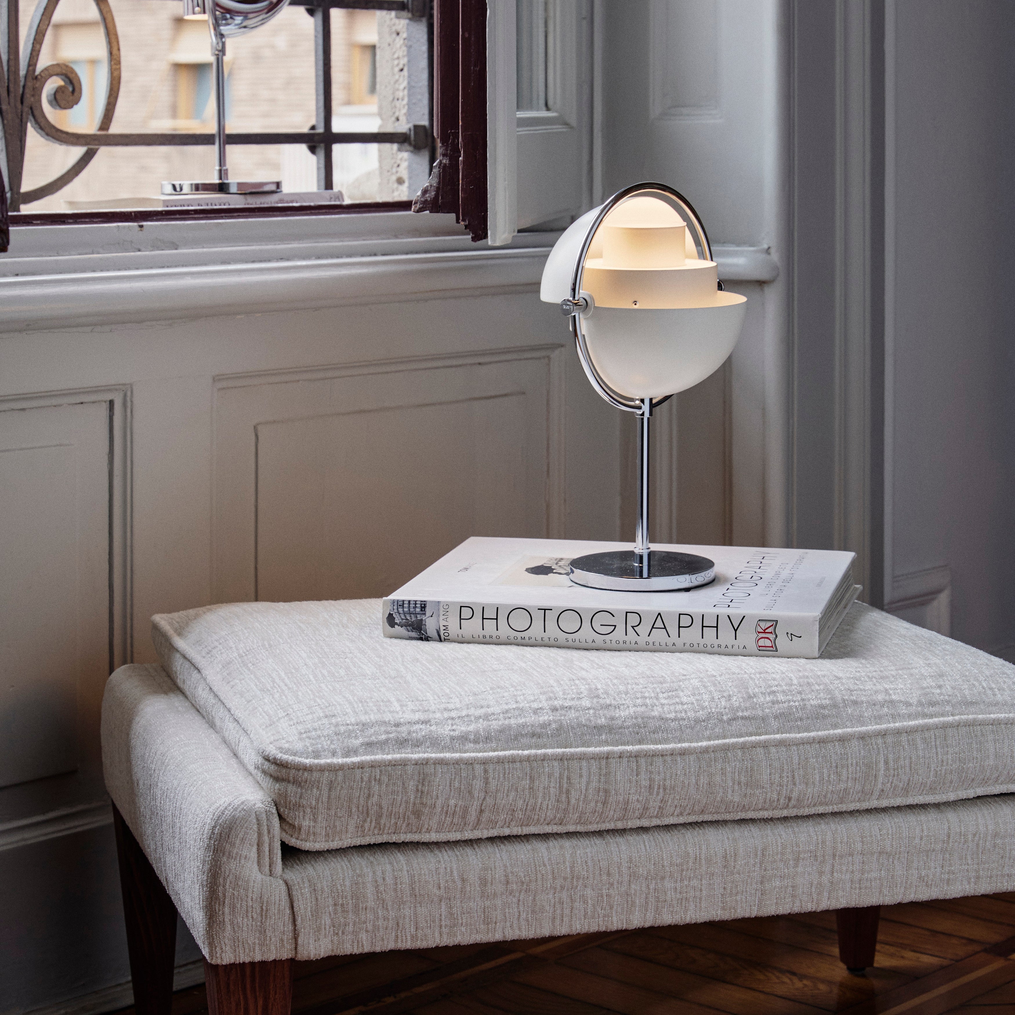 A white half-globe table lamp on a chrome stem and round base, sitting on a book on a cream upholstered bench by a window