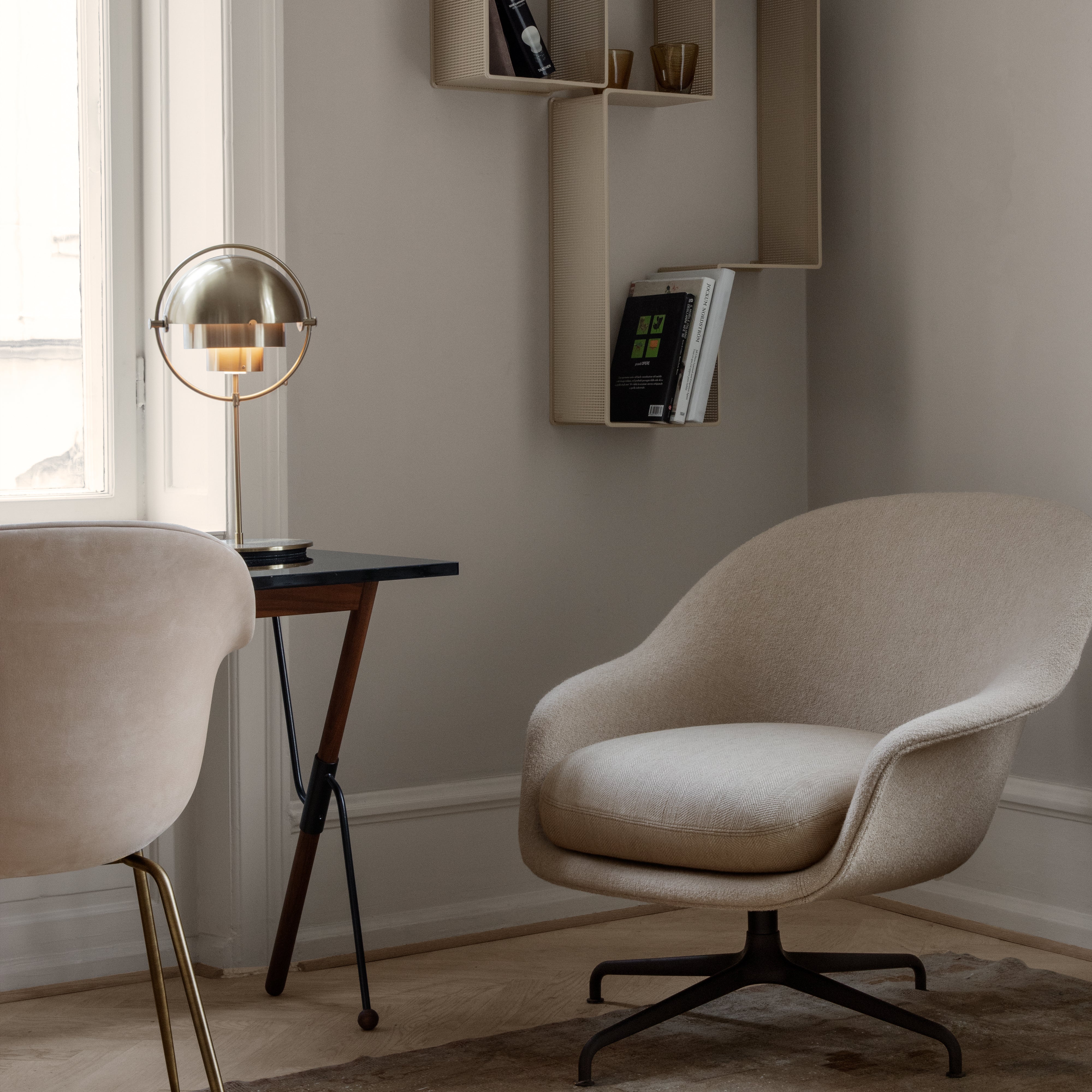 A brass table lamp with a rounded dome shade on a small dark desk beside a cream swivel armchair