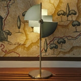 A table lamp with a brass base and stem and pivoting pale green quarter-circle shades, in front of a painted map mural