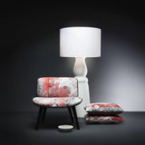A low chair with red and grey patterned upholstery on dark legs beside a tall white lamp and two matching cushions