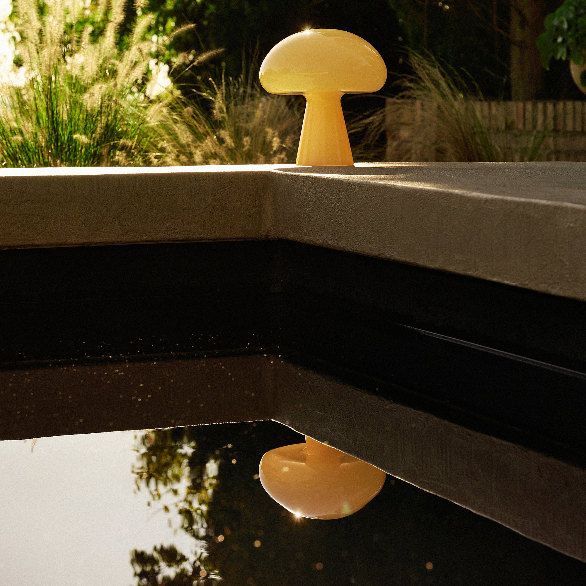 A pale yellow mushroom-shaped glass lamp on a concrete pool edge, reflected in dark water