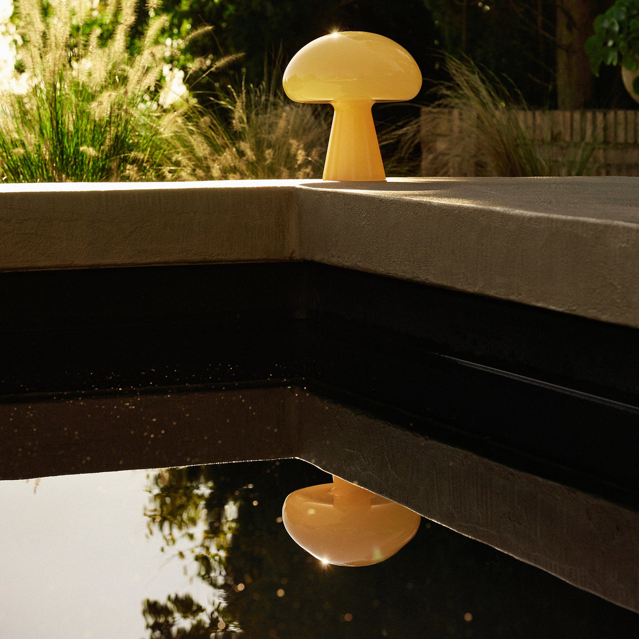 A pale yellow mushroom-shaped glass lamp on a concrete pool edge, reflected in dark water