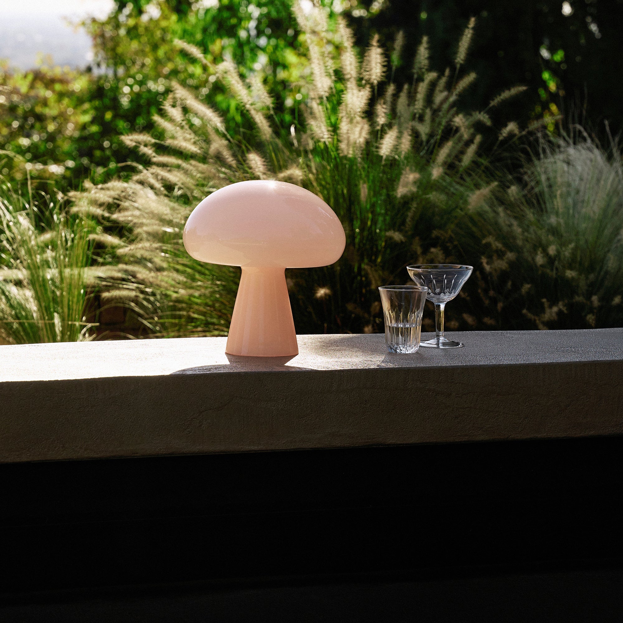 A pale pink glass table lamp shaped like a mushroom on a concrete ledge, with feathery grasses behind