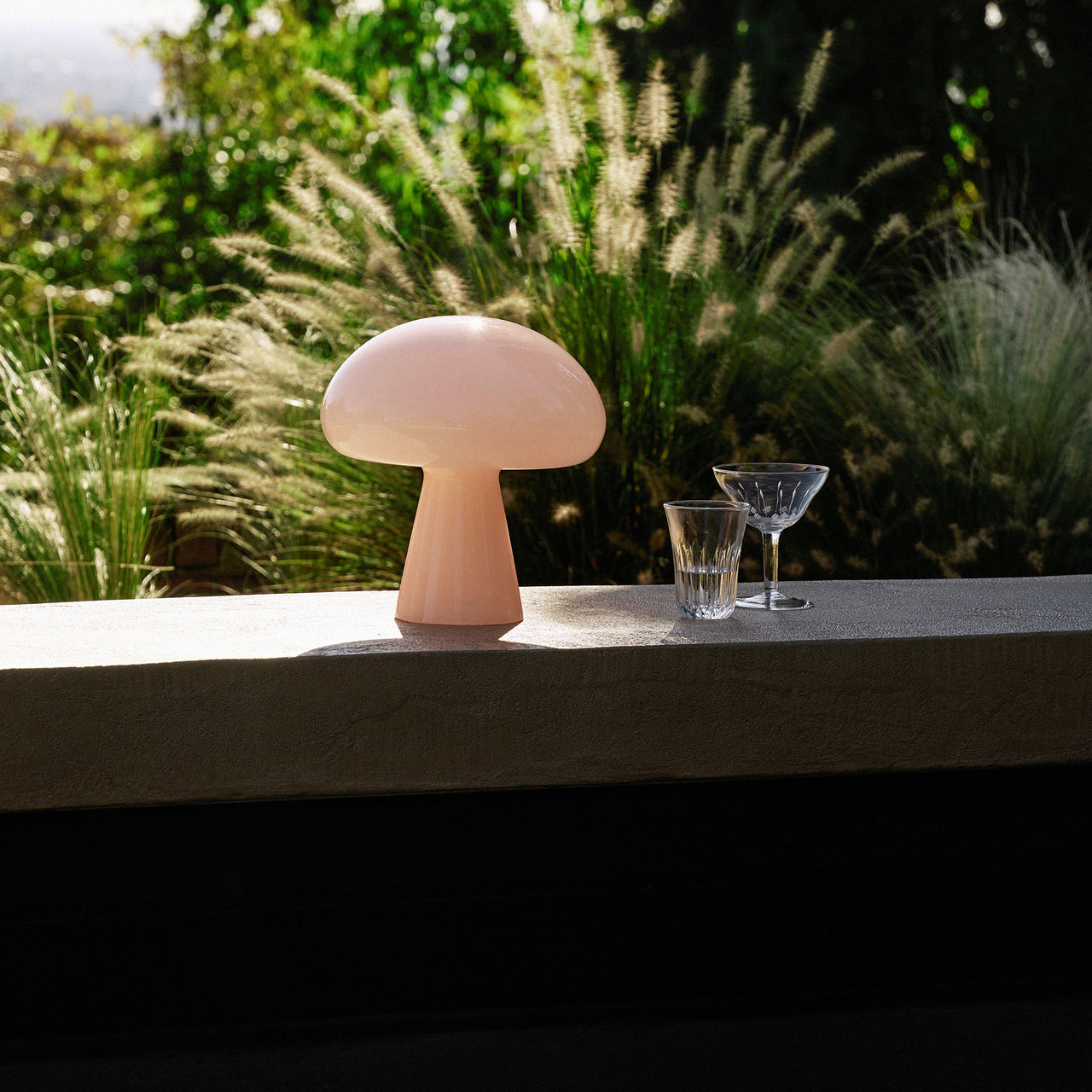 A pale pink glass table lamp shaped like a mushroom on a concrete ledge, with feathery grasses behind