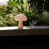 A pale pink glass table lamp shaped like a mushroom on a concrete ledge, with feathery grasses behind