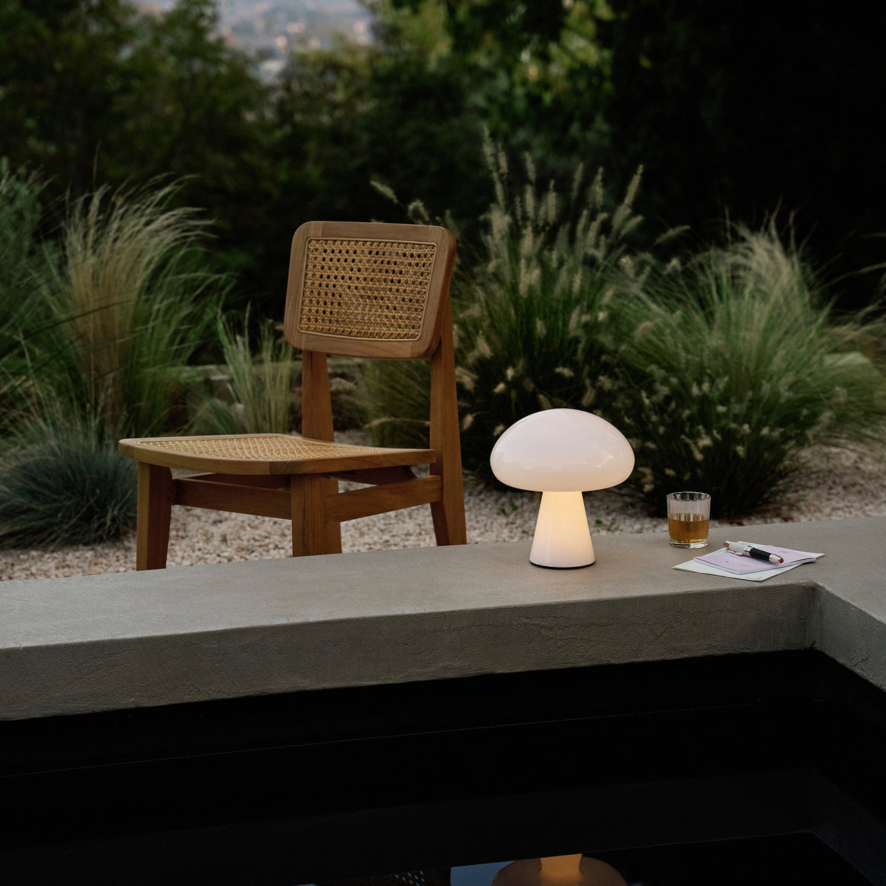 A glowing white mushroom-shaped lamp on a concrete ledge beside a cane-backed wooden chair in a garden at dusk