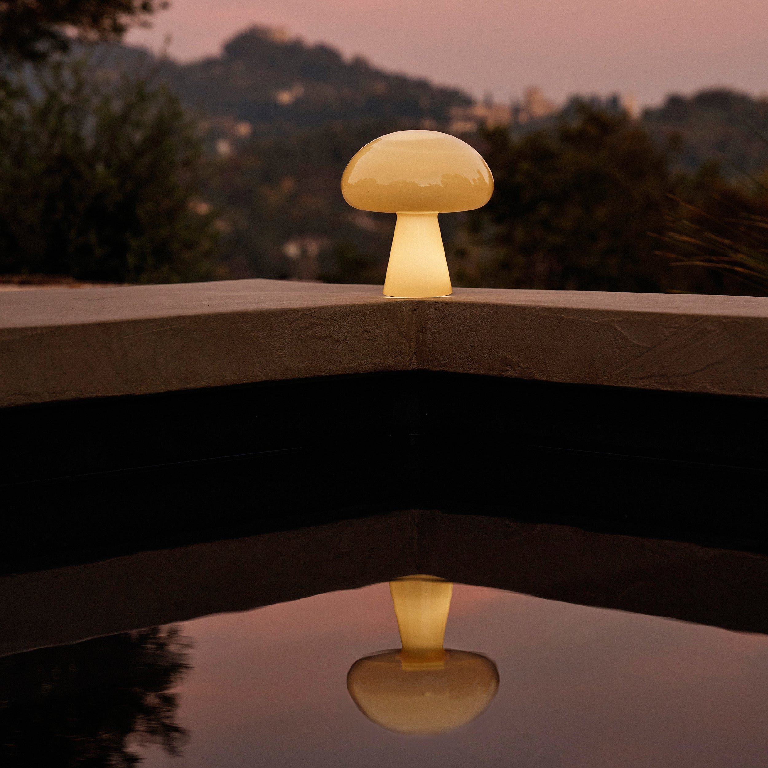 A glowing cream glass lamp with a domed shade on a stone ledge beside a dark pool at dusk
