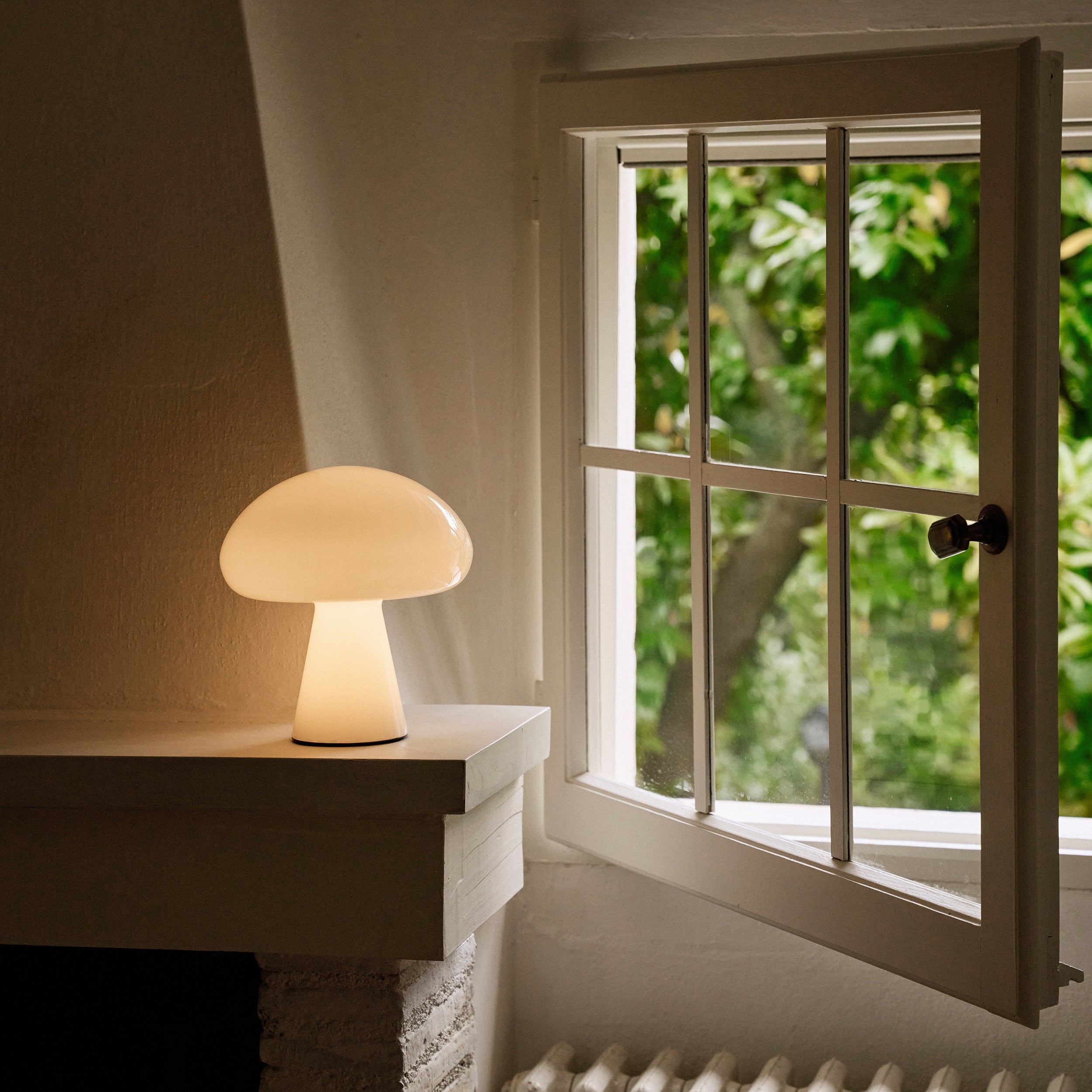 A glowing mushroom-shaped glass lamp on a white mantel beside an open window with green trees outside