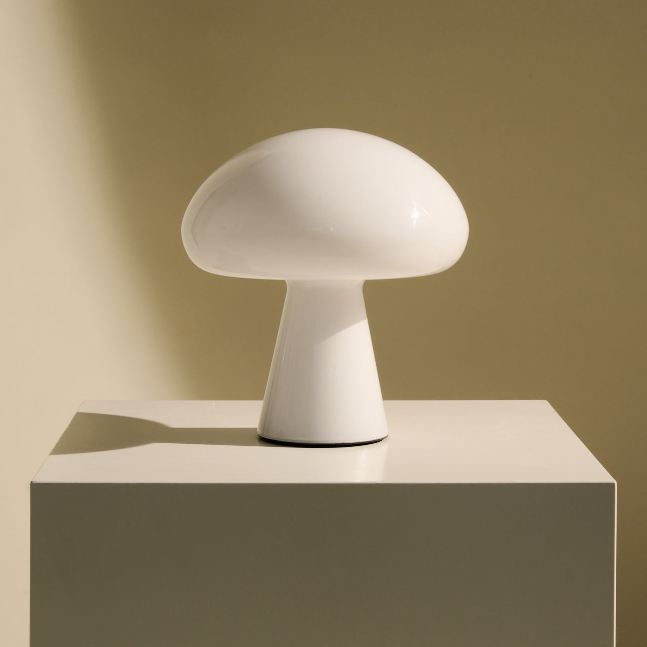 A glossy white glass table lamp shaped like a mushroom, on a cream pedestal against a warm tan wall