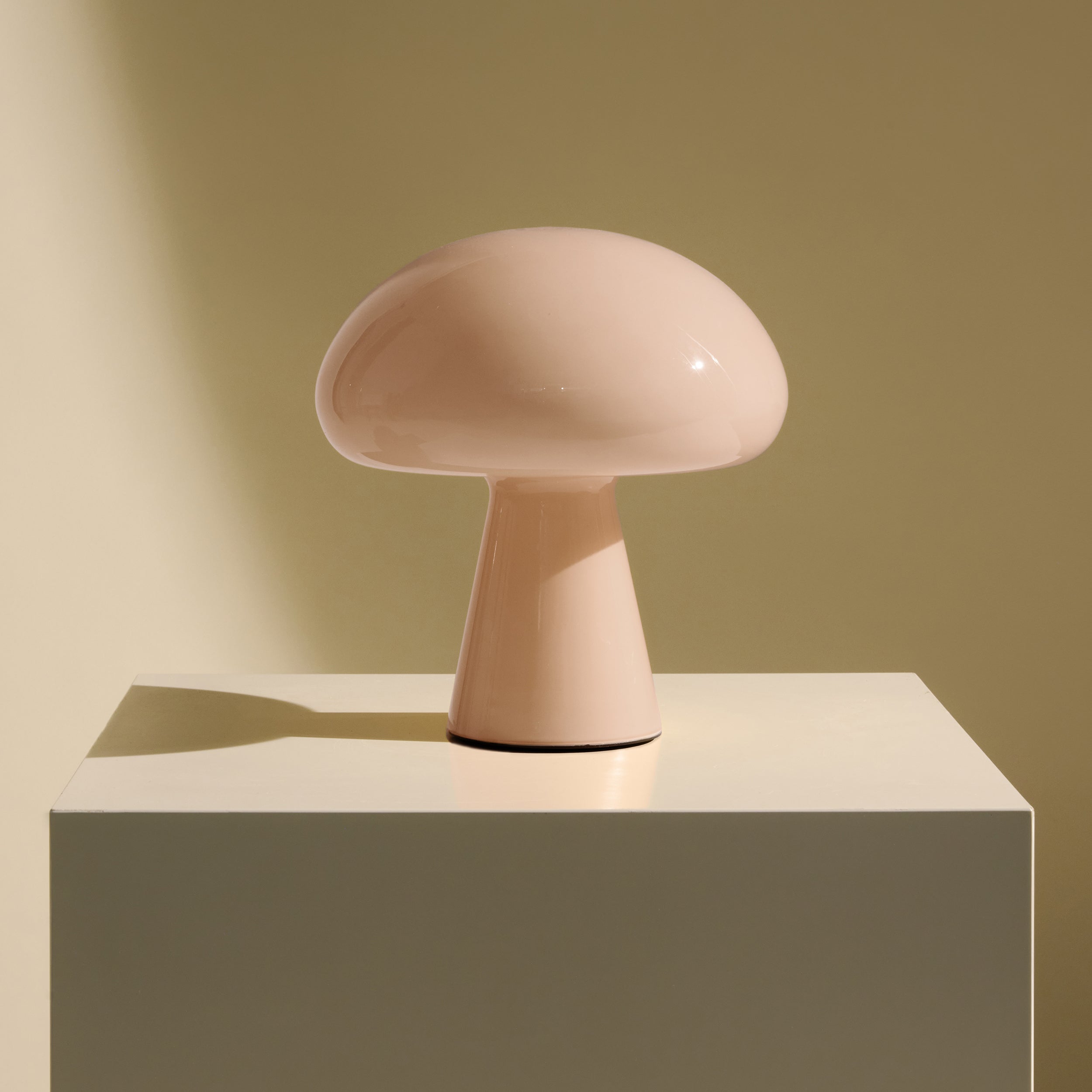 A glossy pale pink table lamp shaped like a mushroom, on a cream pedestal against an olive wall