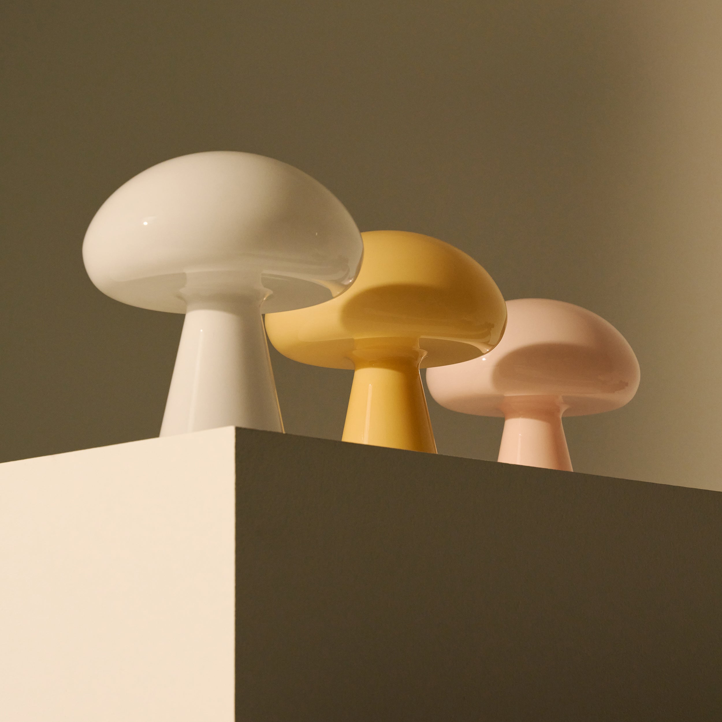 Three glossy glass mushroom-shaped lamps in white, yellow and pale pink on a cream block