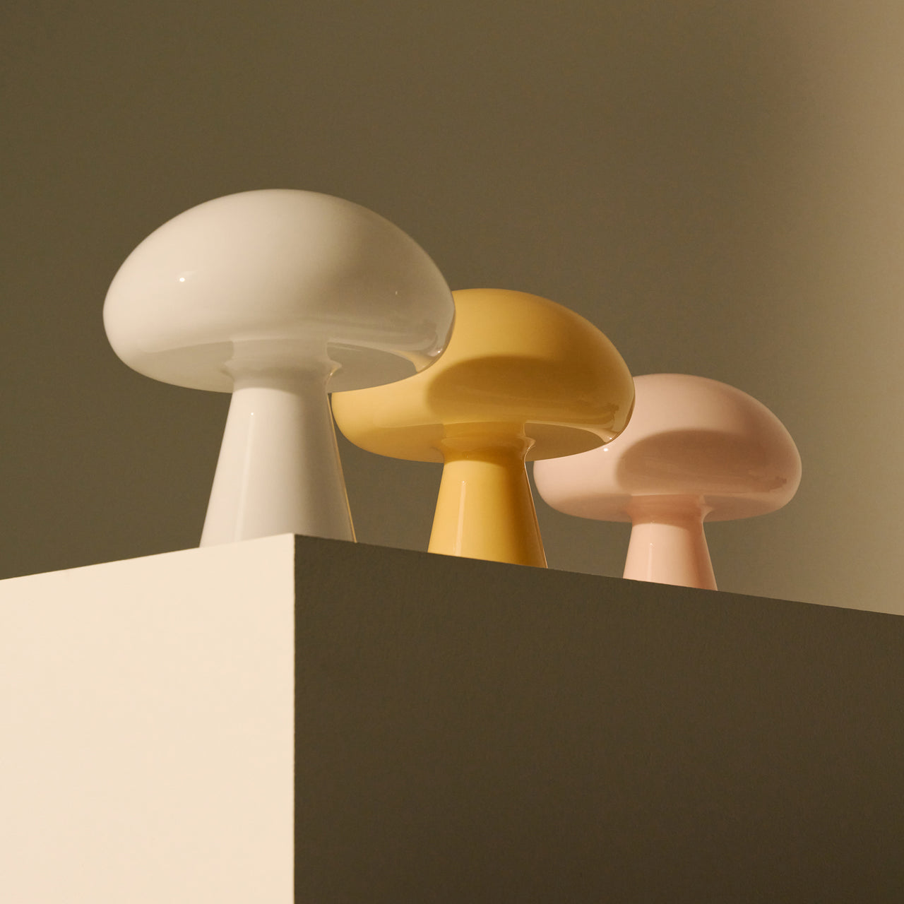 Three glossy glass mushroom-shaped lamps in white, yellow and pale pink on a cream block
