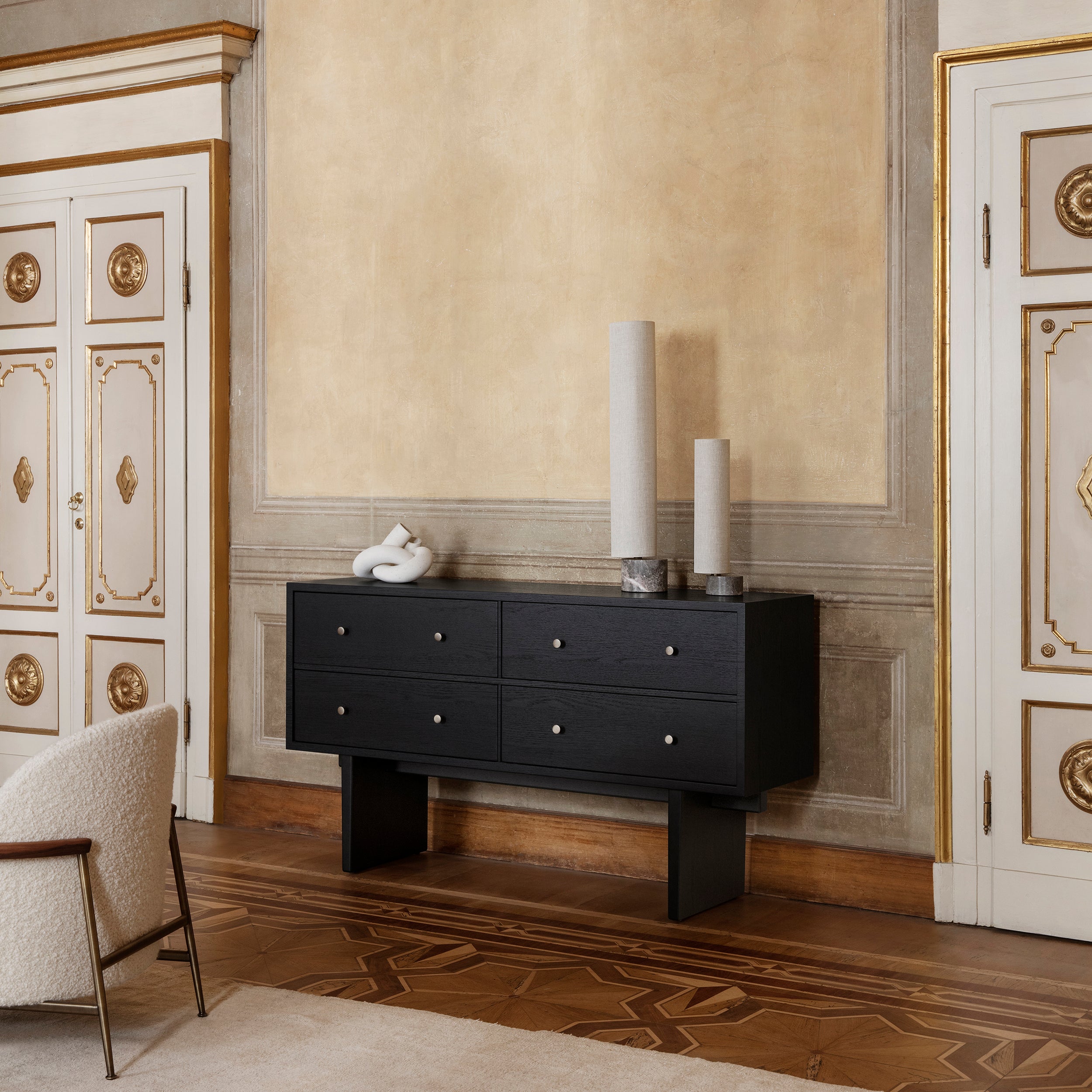 A black sideboard with four drawers and slab legs in an ornate room with gilded doors
